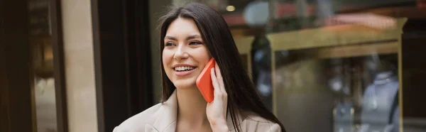 Jovem alegre com cabelos longos morena e maquiagem falando no smartphone com caixa de telefone vermelho e de pé perto da joalheria borrada em Istambul, banner — Fotografia de Stock