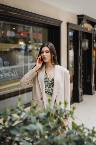 Surprised young woman with brunette long hair and makeup talking on smartphone and standing in trendy outfit with handbag on chain strap near blurred jewelry store and plant in Istanbul — Stock Photo
