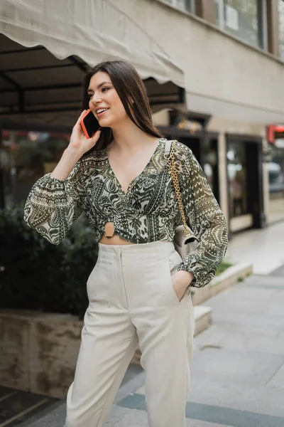 Stylish young woman with brunette long hair and makeup smiling while talking on smartphone and standing in trendy outfit with handbag on chain strap near blurred fancy restaurant in Istanbul — Stock Photo
