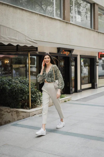 Amazed young woman with brunette long hair and makeup smiling while talking on smartphone and walking in trendy outfit with handbag on chain strap near blurred fancy restaurant in Istanbul — Stock Photo