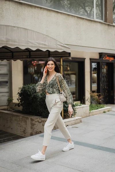 Cheerful young woman with brunette long hair and makeup smiling while talking on smartphone and walking in trendy outfit with handbag on chain strap near blurred fancy restaurant in Istanbul — Stock Photo