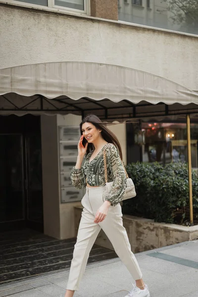 Веселая молодая женщина с длинными волосами и макияжем улыбается, разговаривая по смартфону и прогуливаясь в модном наряде с сумочкой на ремне возле размытого модного ресторана в Стамбуле — стоковое фото