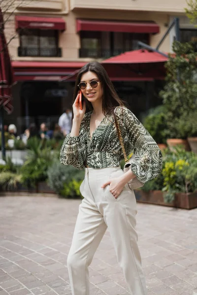 Весела молода жінка з довгим волоссям і сонцезахисними окулярами посміхається під час розмови на смартфоні і стоїть з рукою в кишені і сумочці на ланцюговому ремені біля розмитого будинку і рослин у Стамбулі. — Stock Photo