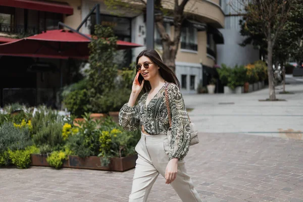 Cheerful young woman with long hair and sunglasses smiling while talking on smartphone and walking in trendy outfit with handbag on chain strap near blurred building and plants on street in Istanbul — Stock Photo