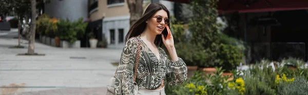 Cheerful young woman with long hair and sunglasses smiling while talking on smartphone and walking with handbag on chain strap near blurred building and plants on street in Istanbul, banner — Stock Photo