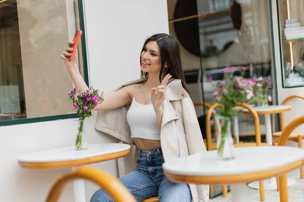 Mujer feliz con el pelo largo sentado en la silla cerca de la mesa del bistro con flores en jarrón y tomar selfie, empujando v signo, posando en ropa de moda en la cafetería en la terraza al aire libre en Estambul - foto de stock