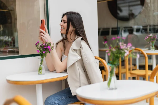 Счастливая женщина с длинными волосами, сидящая на стуле возле бистро стола с цветами в вазе и сообщениями на смартфоне, сидя в модной одежде в кафе на террасе в Стамбуле — стоковое фото