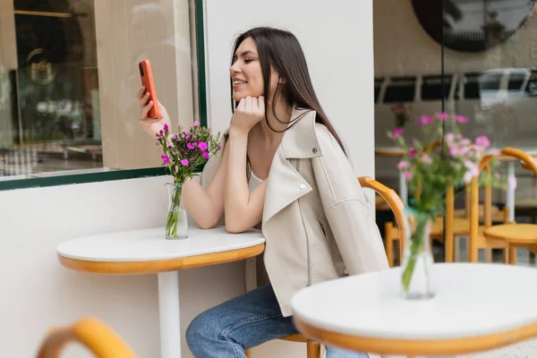 Mujer feliz con el pelo largo sentado en la silla cerca de la mesa del bistro con flores en jarrón y mensajes de texto en el teléfono inteligente mientras está sentado en ropa de moda con chaqueta beige en la cafetería en la terraza al aire libre en Estambul - foto de stock