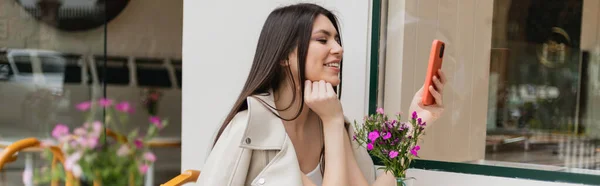Happy woman with long hair sitting near blurred flowers and texting on smartphone while sitting in trendy clothes with beige leather jacket in cafe on terrace outdoors in Istanbul, banner — Stock Photo