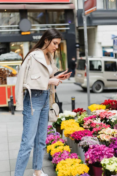 Femme brune aux cheveux longs debout en veste en cuir beige, jean denim et sac à main avec bracelet chaîne tenant smartphone tout en regardant des fleurs près de voitures floues dans la rue à Istanbul, vendeur — Photo de stock