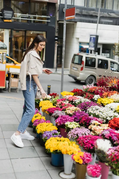 Happy woman with long hair standing in beige leather jacket, denim jeans and handbag with chain strap, holding smartphone near bouquet of flowers next to blurred cars on street in Istanbul, vendor — Stock Photo