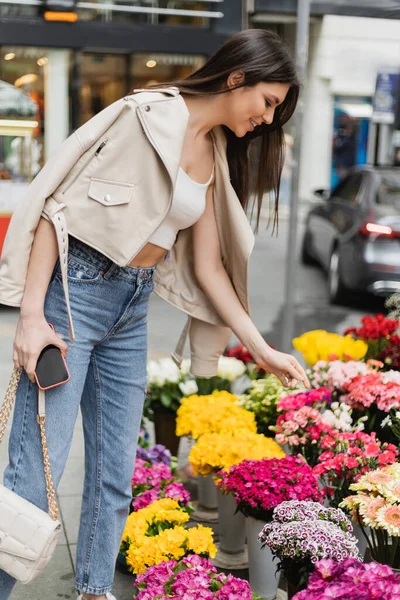 Весела молода жінка з довгим волоссям стоїть у бежевій шкіряній куртці, тримаючи сумочку з ланцюговим ременем і смартфоном біля квітів на вулиці Стамбула, постачальник — Stock Photo