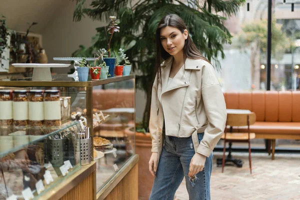 Brunette young woman with long hair in beige leather jacket and denim jeans looking at camera while standing near cake display with pastry and jars of jam in modern bakery shop in Istanbul — Stock Photo