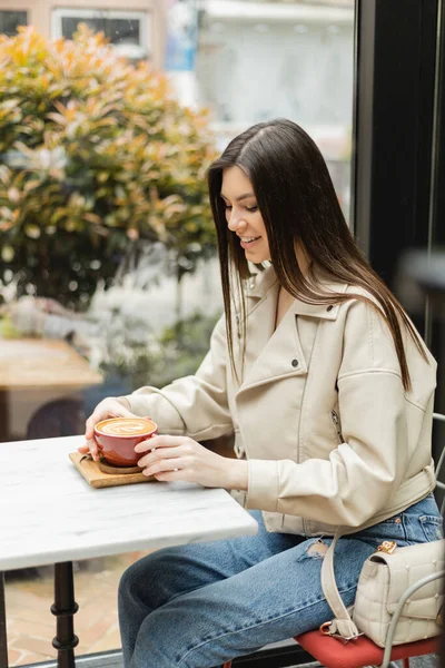 Glückliche brünette Frau in Lederjacke sitzt auf einem Stuhl neben Fenster und Bistrotisch und hält eine Tasse Cappuccino mit Kaffeekunst in einem modernen Café in Istanbul — Stockfoto
