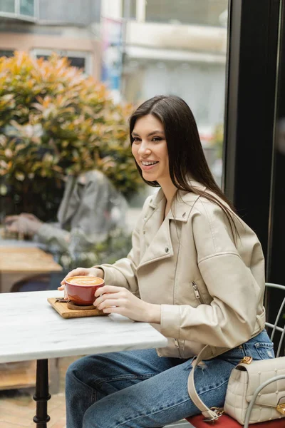 Glückliche brünette Frau in Lederjacke sitzt auf einem Stuhl neben Fenster und Bistrotisch und hält eine Tasse leckeren Cappuccino mit Kaffeekunst in einem modernen Café in Istanbul — Stockfoto
