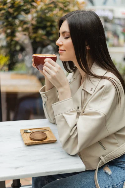 Happy brunette woman with closed eyes in leather jacket sitting on chair next to window and bistro table while holding cup of cappuccino inside of modern cafe in Istanbul — Stock Photo