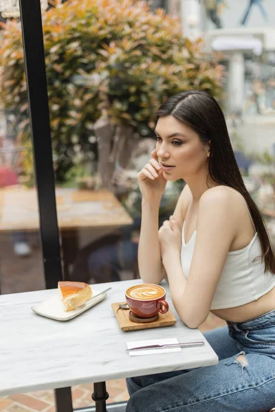 Mujer joven con el pelo largo sentado junto a la ventana y mesa de bistró con taza de capuchino y sabroso pastel de queso en el plato mientras mira hacia el interior de la cafetería moderna en Estambul - foto de stock