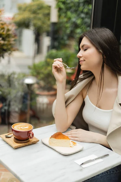 Jeune femme aux cheveux longs tenant fourchette près du gâteau au fromage à côté de la tasse de cappuccino sur la table de bistrot tout en étant assis dans une veste en cuir près de la fenêtre à l'intérieur du café moderne à Istanbul — Photo de stock