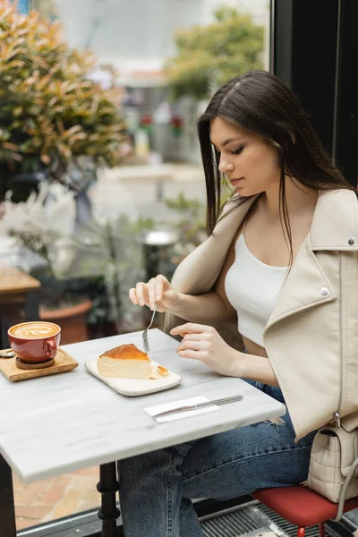 Jeune femme brune tenant fourchette près du gâteau au fromage à côté de la tasse de cappuccino avec art du café sur la table de bistrot tout en étant assis dans une veste en cuir près de la fenêtre à l'intérieur du café moderne à Istanbul — Photo de stock