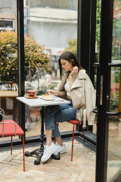 Полная длина молодая женщина с длинными волосами держа вилку возле чизкейка и чашку капучино на бистро стол, сидя в кожаной куртке рядом окно внутри современного кафе в Стамбуле — стоковое фото