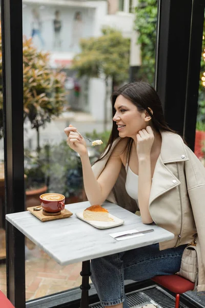 Femme gaie avec les cheveux longs tenant fourchette près cheesecake à côté de tasse de cappuccino avec art du café sur la table de bistrot tout en étant assis dans une veste en cuir près de la fenêtre à l'intérieur du café moderne à Istanbul — Photo de stock