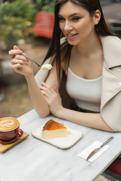 Femme souriante aux cheveux longs tenant fourchette avec gâteau au fromage à côté de la tasse de cappuccino avec art du café sur la table de bistrot tout en étant assis à l'intérieur du café moderne à Istanbul — Photo de stock