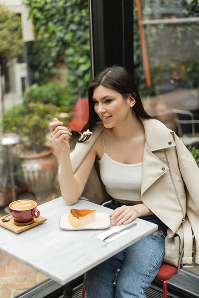 Femme souriante aux cheveux longs tenant fourchette près cheesecake à côté de tasse de cappuccino avec art du café sur la table de bistrot tout en étant assis à côté de la fenêtre à l'intérieur du café moderne à Istanbul — Photo de stock