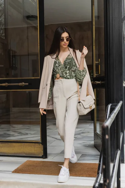 Brunette and young woman in trendy outfit and sunglasses walking out of building while holding handbag with chain strap on modern street in Istanbul, entrance door — Stock Photo