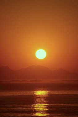 Arka planda dağlar ve önünde deniz olan gün batımı. turuncu gökyüzü. Huzur ve yansıma