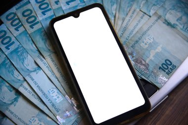 Beyaz ekranlı cep telefonu ve Brezilya para birimi (REAL) arka planda. Başvuru, ekonomi, yatırım ve Brezilya para birimi işlemleri kavramı.