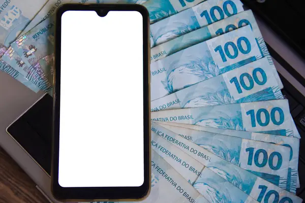 Beyaz ekranlı cep telefonu ve Brezilya para birimi (REAL) arka planda. Başvuru, ekonomi, yatırım ve Brezilya para birimi işlemleri kavramı.