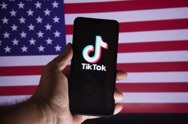 Elinde TIKTOK uygulama logosu ve arka planda ABD bayrağı olan bir akıllı telefon var. Ekranında tiktok uygulaması olan cep telefonu. Rio de Janeiro, Brezilya - 01.16.2025.