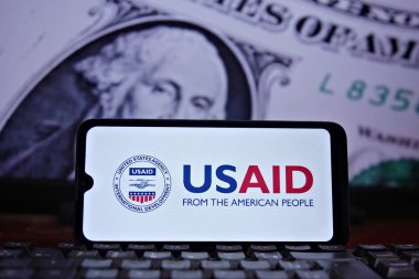 Ön planda ABD Uluslararası Kalkınma Dairesi (USAID) mührü ve arka planda ABD bayrağı ve doları bulunan cep telefonu. USAID. Rio de Janeiro, Brezilya - 02.04.2025