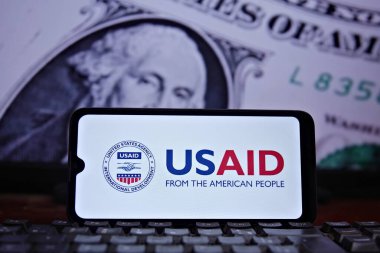 Cell phone with the United States Agency for International Development (USAID) seal in the foreground and the US flag and dollar in the background. USAID. Rio de Janeiro, Brazil - 02.04.2025 clipart