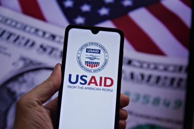 Cell phone with the United States Agency for International Development (USAID) seal in the foreground and the US flag and dollar in the background. USAID. Rio de Janeiro, Brazil - 02.04.2025 clipart