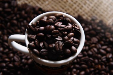 Kahve fincanında kavrulmuş ve kahverengi kahve çekirdekleri. Üst Manzara. Kahve torbasıyla birlikte rustik bir yüzeyde kavrulmuş kahve çekirdekleri. Yan ışık