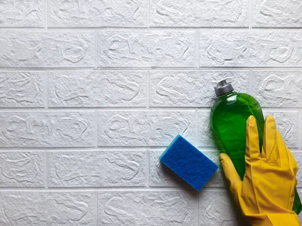 Concept housekeeping spring cleaning. Copy space. Flat lay Hand in yellow rubber glove holds bottle of green detergent, next to it lies blue kitchen cleaning sponge on white brick wall background. 