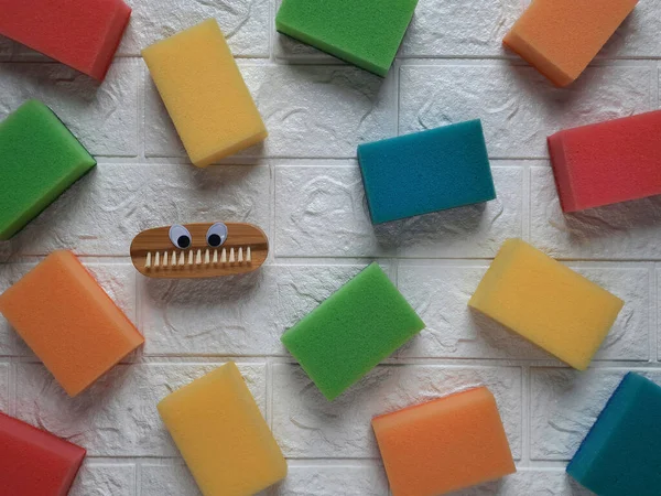 Background from kitchen sponges. In center is cleaning brush with eyes. Housecleaning concept.Multi colored sponges for cleaning and wooden brush are scattered on white wall. Flat lay.