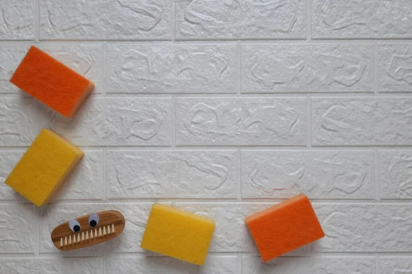 Fun brush and kitchen sponges for cleaning. Copy space. Fun cleaning concept.  Scrubbing brush with eyes and dishwashing sponge on brick wall background. Flat lay. Household duties for children.