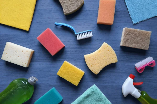 Background with items for cleaning house. Flat lay. Kitchen sponges of various shapes, rags, brushes, dish detergent, cleaning spray lie on dark blue wooden table. Hygiene concept. Top view.
