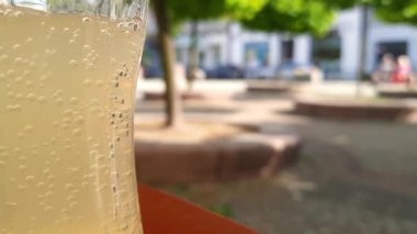 Tahta masada bir bardak meşrubat. Arka planda bulanık yaz güneşli şehir manzarası. Soğuk içecekteki gaz baloncuklarını yakın çekim. Su dengesi teması, susuzluk, yaz sıcağı, şehir molası