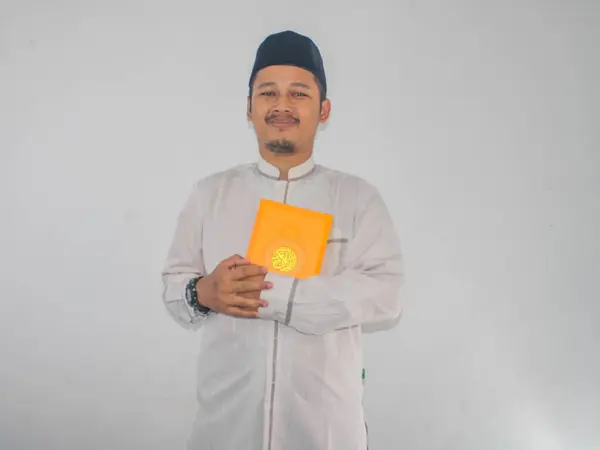 Müslüman Asyalı bir adam elinde Kuran tutarken kameraya gülümsüyor.