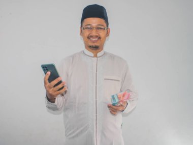 Müslüman Asyalı adam cep telefonu ve para tutarken mutlu gülümsüyor