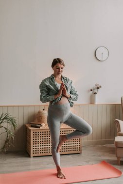 Rahat kıyafetler giyen hamile bir kadının evde oturma odasında tek ayak üzerinde yoga yaparken tam dikey görüntüsü. İnsanlar ve hamilelik aktiviteleri konsepti. Sağlıklı ebeveyn kavramı. 