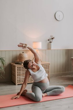 Mutlu, gülümseyen, hamile bir kadının sabah evde paspasa uzanmasının en güzel görüntüsü. Bekleyen genç anne yoga egzersizi yapıyor. Spor giyim sektöründe esnek bir kadın egzersiz yapıyor.. 