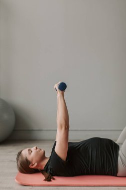 Oturma odasında paspasın üzerinde duran dambıllarla egzersiz yapan genç hamile bir kadın. Hamile kadın konsepti için spor. Sağlıklı hamilelik. 4 bin. Yüksek kalite 4k görüntü