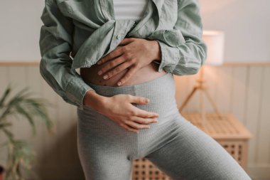 Rahat kıyafetler giyen hamile bir kadının evde oturma odasında tek ayak üzerinde yoga yaparken yakın plan fotoğrafı. İnsanlar ve hamilelik aktiviteleri konsepti. Sağlıklı ebeveyn kavramı. 