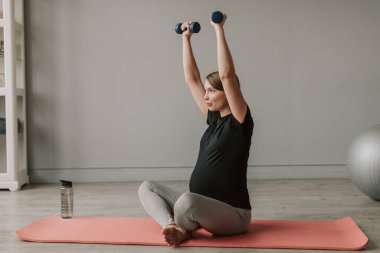 Spor giyimli, mutlu ve hamile bir kadın oturma odasında egzersiz yapıyor. Hamilelik sırasında doğum öncesi bakımı. Hamilelik süresince yaşam tarzı, sağlık ve spor kavramlarını.