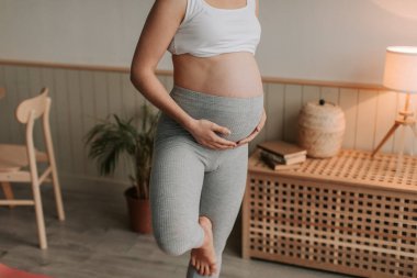 Rahat kıyafetler giyen hamile bir kadının evde oturma odasında tek ayak üzerinde yoga yaparken yakın plan fotoğrafı. İnsanlar ve hamilelik aktiviteleri konsepti. Sağlıklı ebeveyn kavramı.