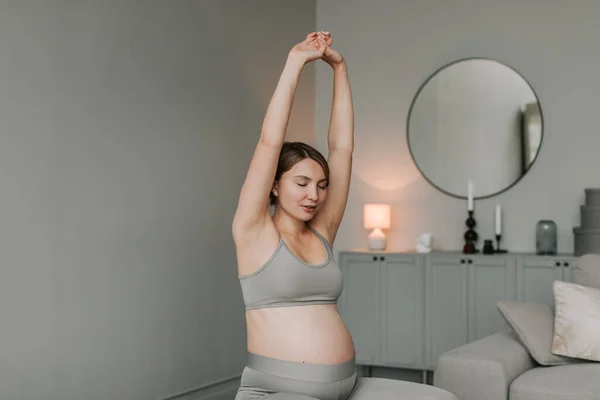 Full shot of young expectant woman with big belly in sportswear doing breathing and stretching exercises sitting on fitness ball in living room at home. Healthy Pregnancy concept